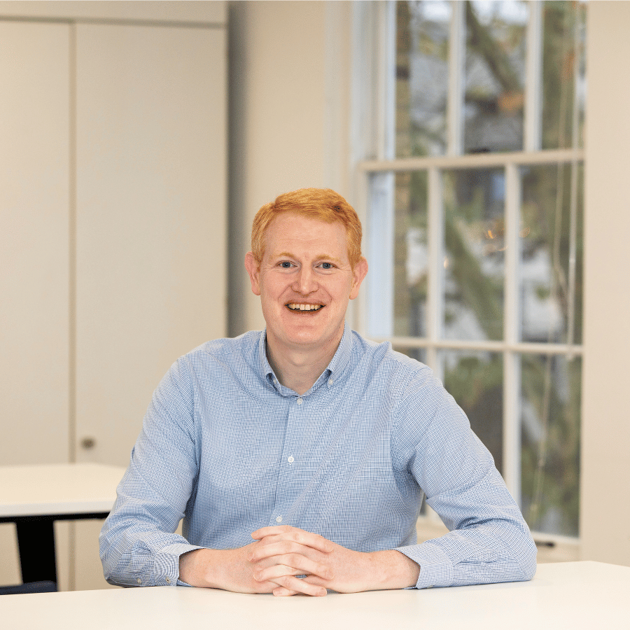 Man wearing a light blue patterned shirt seated at a white table in a bright room with a large window.