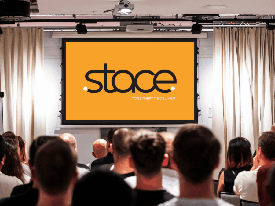 Group of people sitting on chairs in front a projector screen displaying the Stace company logo.