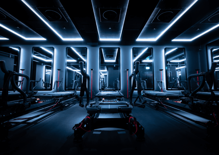 A dark, modern gym room with multiple treadmills and illuminated ceiling and wall lights reflecting off mirrors.