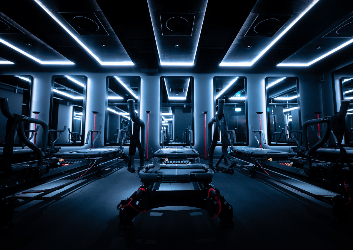 A dark, modern gym room with multiple treadmills and illuminated ceiling and wall lights reflecting off mirrors.