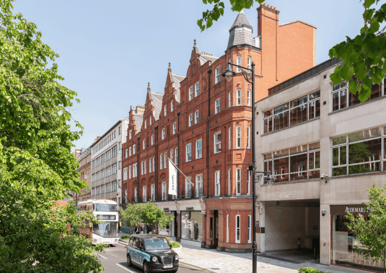 A street view featuring a red-brick Victorian building, modern offices, a double-decker bus, a black taxi, and leafy trees under a clear sky.