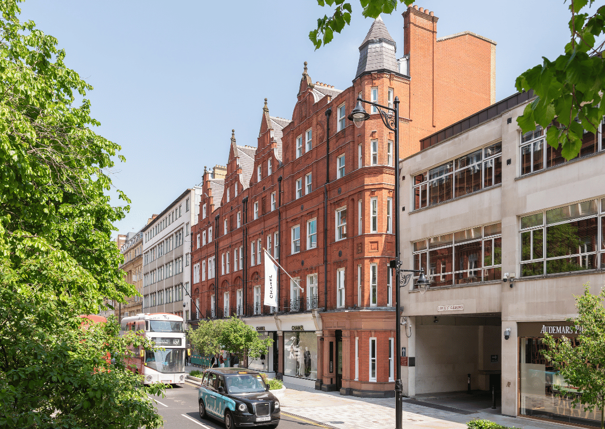 A street view featuring a red-brick Victorian building, modern offices, a double-decker bus, a black taxi, and leafy trees under a clear sky.