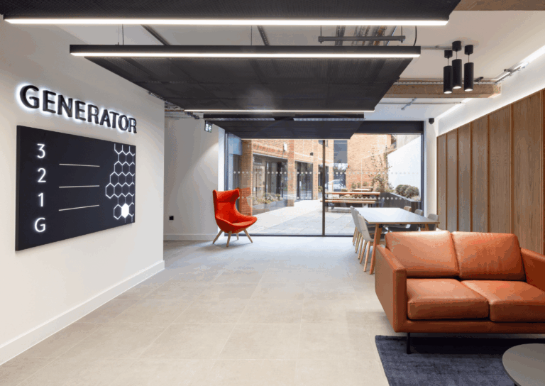 Modern office lounge featuring a burnt orange sofa, red high-back chair, and long communal table with chairs.