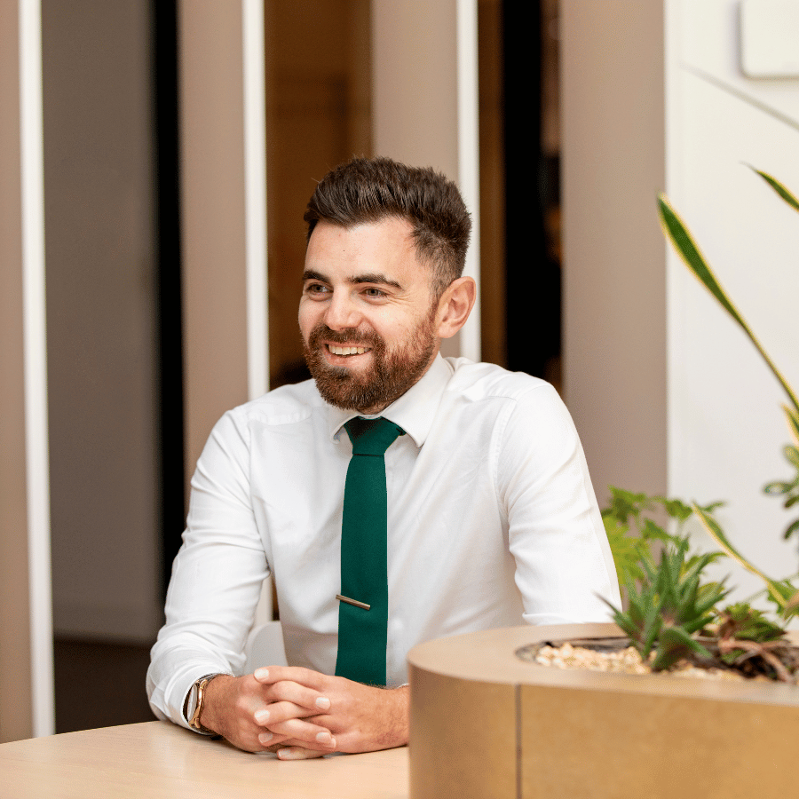Man wearing a white shirt and green tie seated at a table with plants in the foreground.