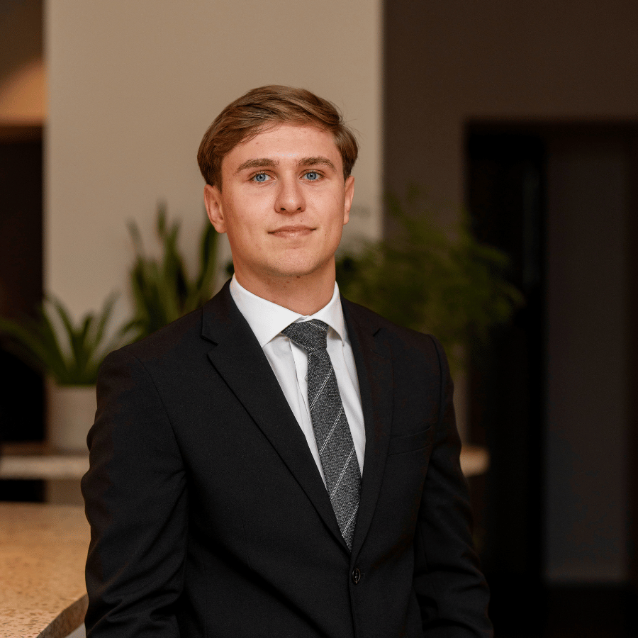 Man wearing a black suit with a white shirt and patterned tie, standing indoors near a countertop with green plants in the background.