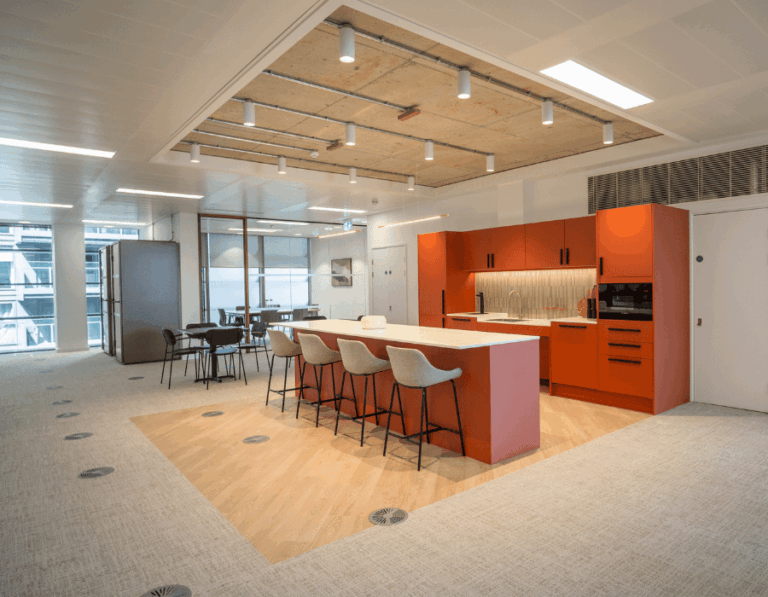 Modern office space with a wooden table, lounge area, and a kitchen counter with orange cabinets and bar stools.