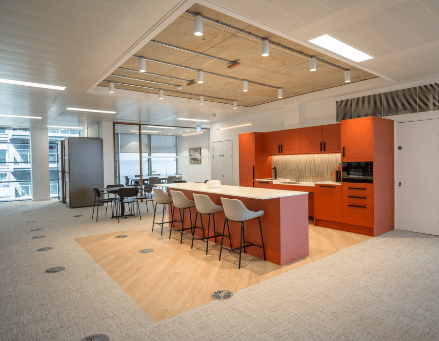 Modern office space with a wooden table, lounge area, and a kitchen counter with orange cabinets and bar stools.