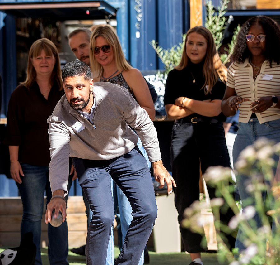 Man about to throw a petanque ball, looked on by a group of five others