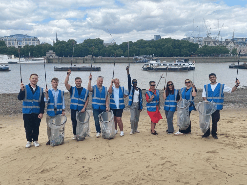 A group of people wearing blue safety vests standing on a sandy riverbank, holding litter pickers and clear bags filled with collected waste.