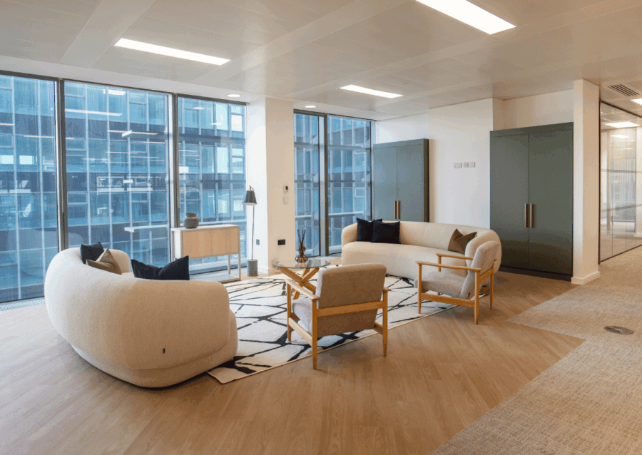 Modern office lounge with two curved sofas, two wooden chairs, and a patterned rug near large glass windows.