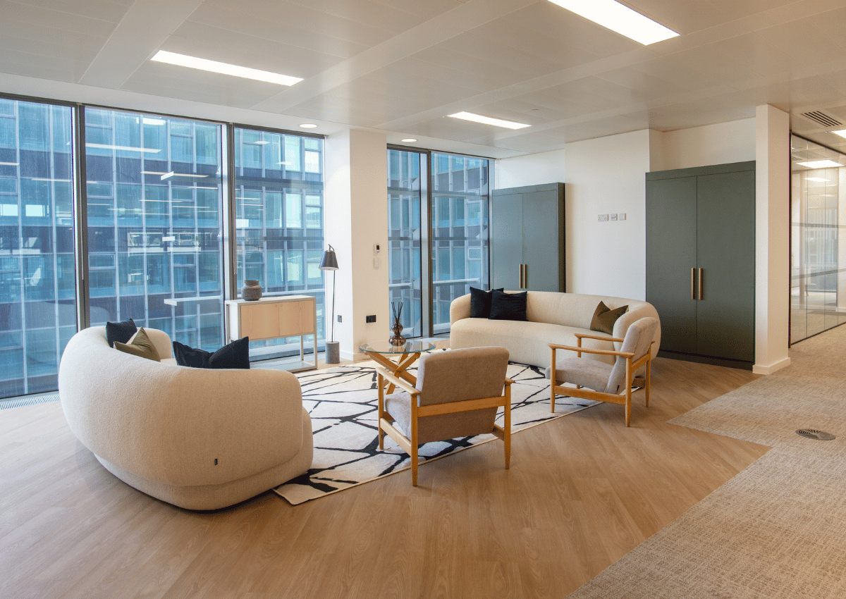 Modern office lounge with two curved sofas, two wooden chairs, and a patterned rug near large glass windows.
