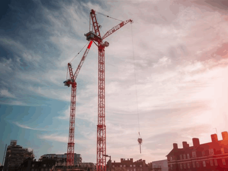 Two tall red construction cranes against a partly cloudy sky with buildings in the foreground.