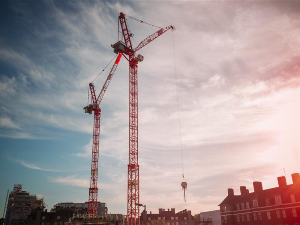 Two tall red construction cranes against a partly cloudy sky with buildings in the foreground.